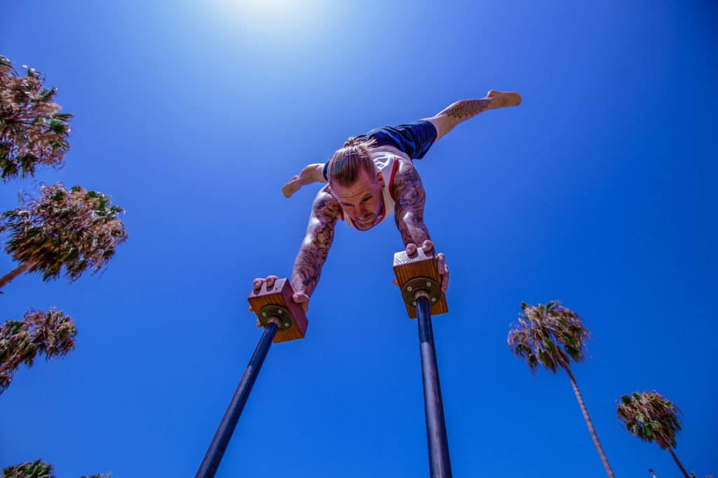 Bottom View of Coach Kyle Weiger doing half straddle handstand on a H-shape Canes Trainer with Tropical Trees Background