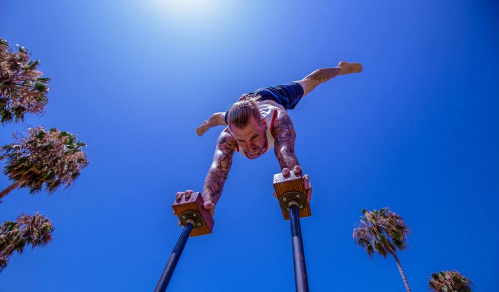 Bottom View of Coach Kyle Weiger doing half straddle handstand on a H-shape Canes Trainer with Tropical Trees Background