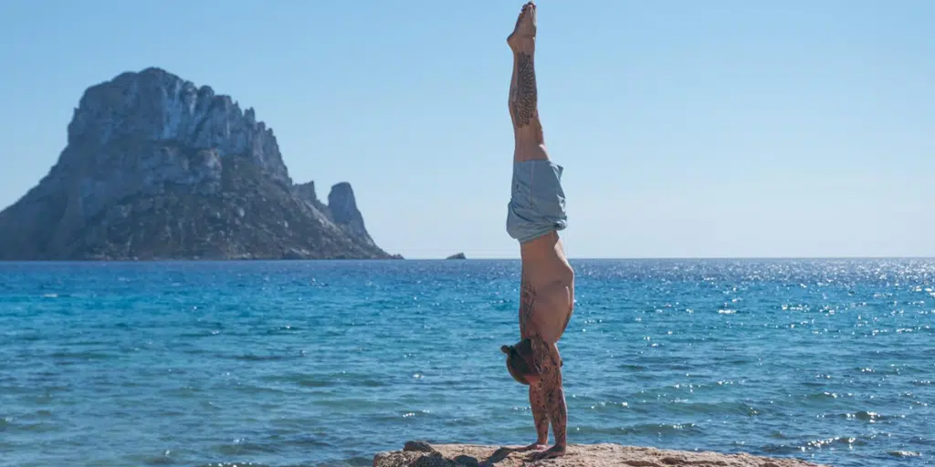 Side view of coach Kyle Weiger handstanding at the side of the ocean with a scenic view of an island mountain