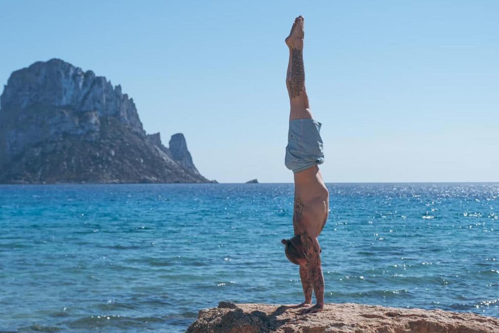 Side view of coach Kyle Weiger handstanding at the side of the ocean with a scenic view of a rocky island mountain