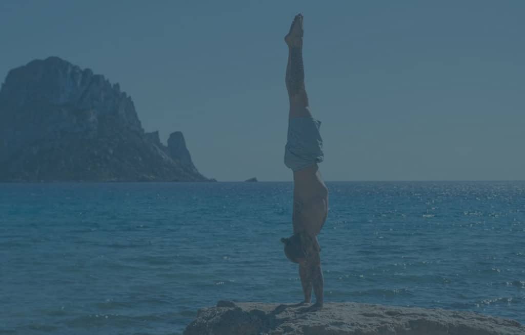 Side view of coach Kyle Weiger handstanding on a rock next to the ocean with a view of a small rocky island with teal filter