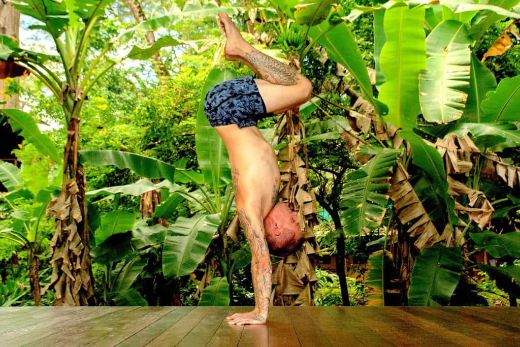 Coach Kyle Weiger doing a tucked handstand on a wooden floor with a tropical background