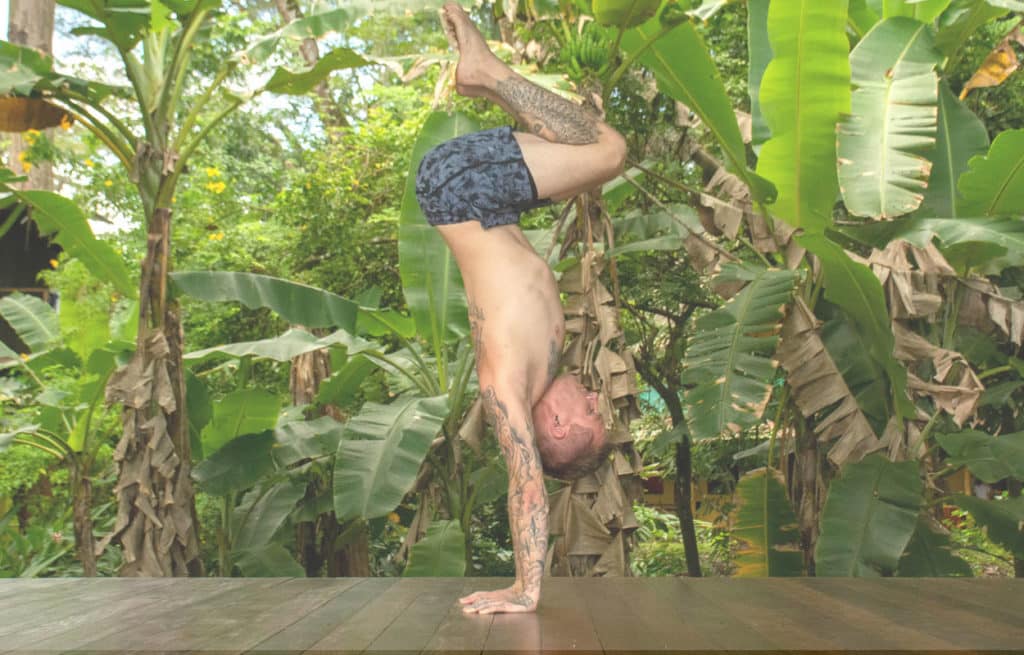 Coach Kyle Weiger doing a tucked handstand on a wooden floor with a tropical background and white filter