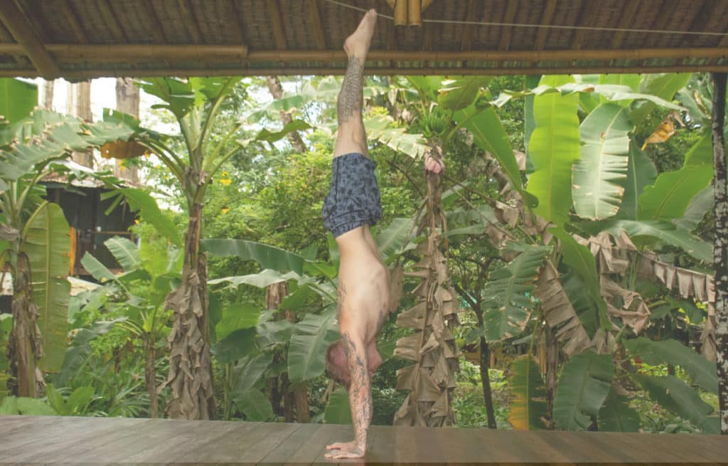 Coach Kyle Weiger doing a handstand on a wooden floor with a tropical background and white filter