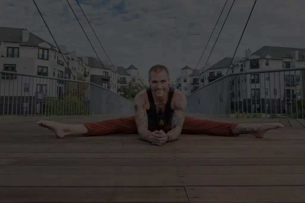 Smiling handstand coach Kyle Weiger front view splitting on wooden floor with houses in the background with dark teal filter