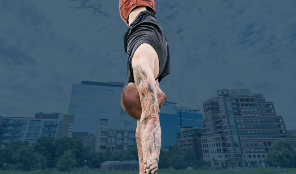 Side view of coach Kyle Weiger handstanding on the grass with darkened buildings and cloudy sky background