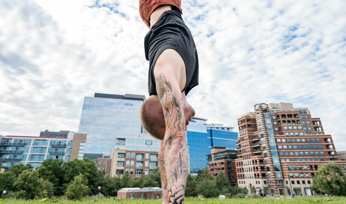 Side view of coach Kyle Weiger handstanding on the grass with darkened buildings and cloudy sky background