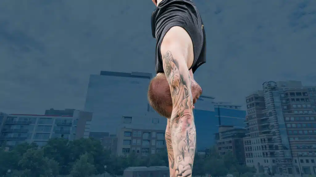 Side view of coach Kyle Weiger handstanding on the grass with darkened buildings and cloudy sky with dark teal background